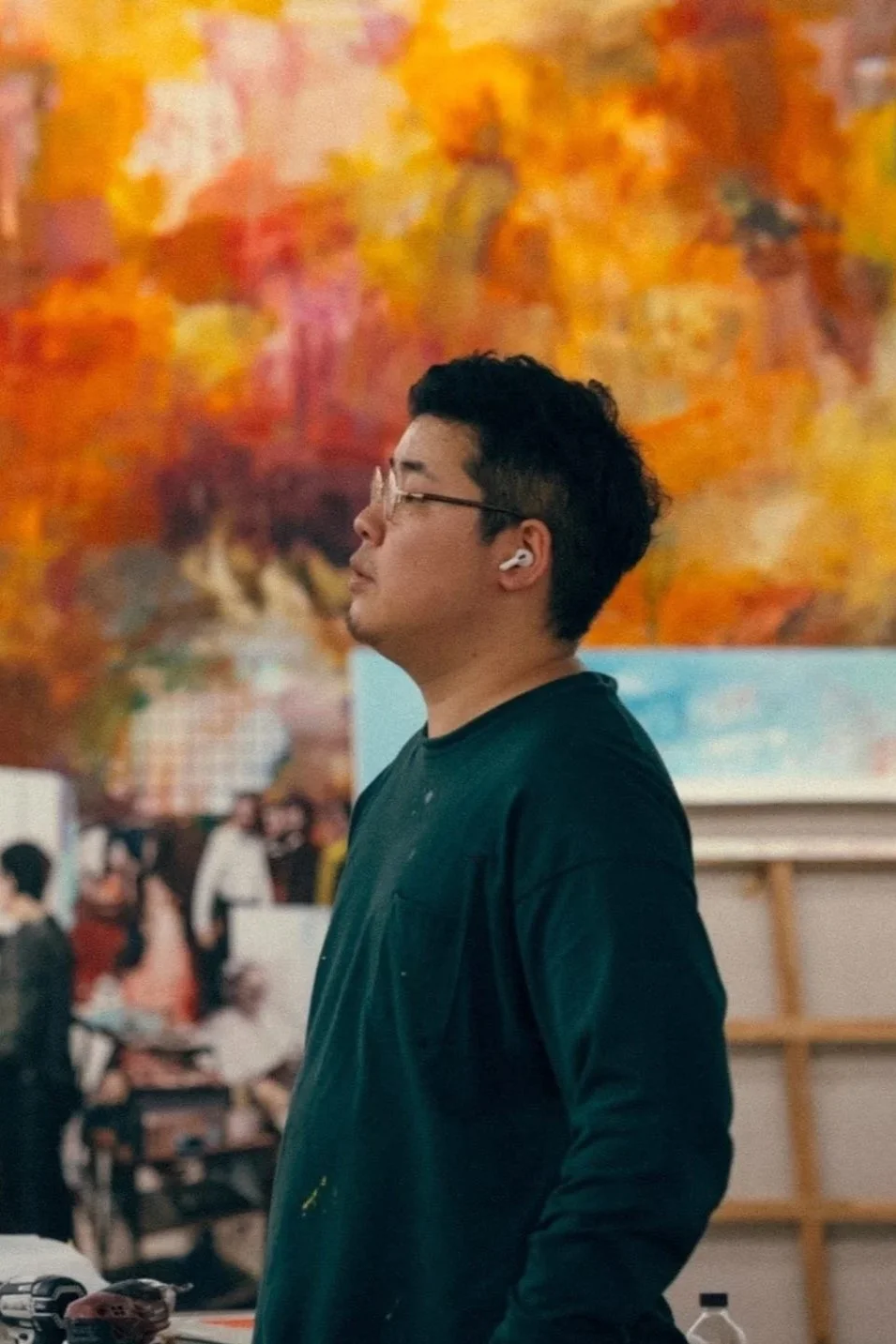 A man with glasses, wearing earbuds and a dark shirt, stands in profile in front of an abstract orange and red background at an art gallery or event.