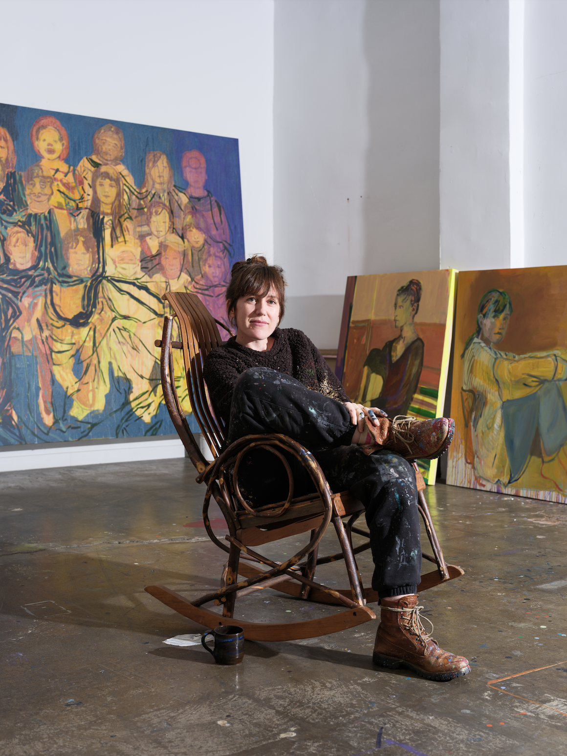 Female artist sitting on a wooden rocking chair in her studio, surrounded by large colorful paintings, some depicting portraits of women and others with abstract and expressive art styles.