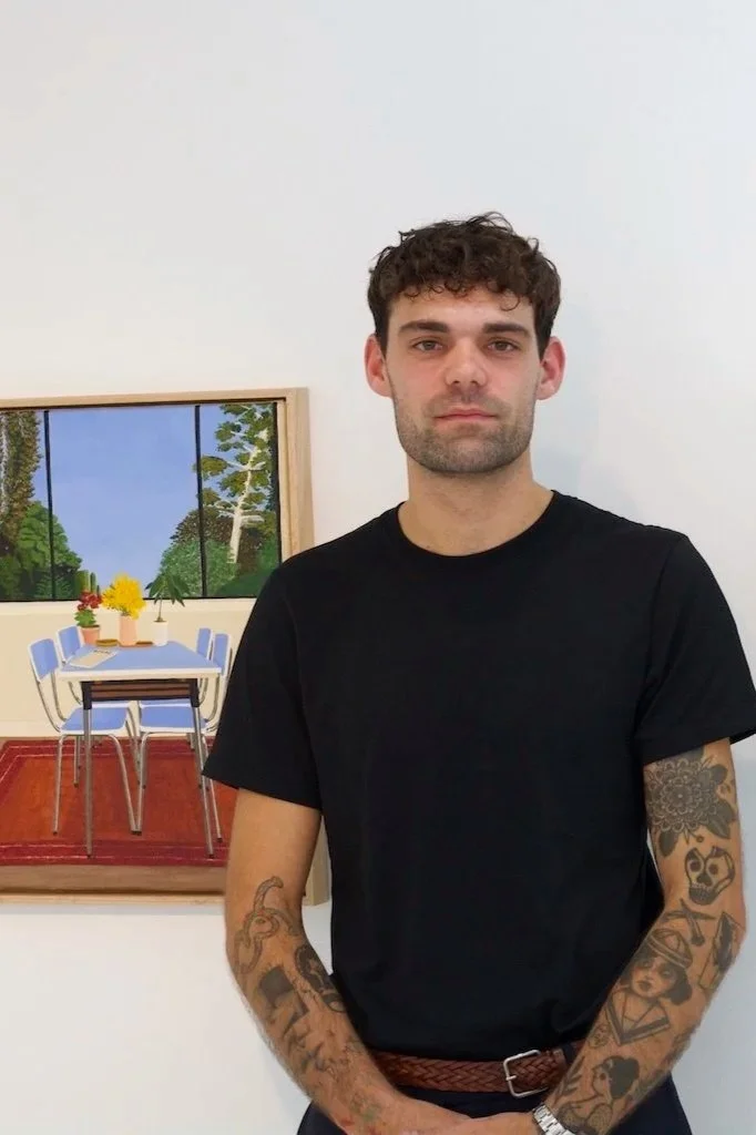 A young man with curly dark hair and tattoos on his arms, wearing a black t-shirt, standing in front of a colorful painting of a dining room with a table, chairs, and a window showing trees outside.