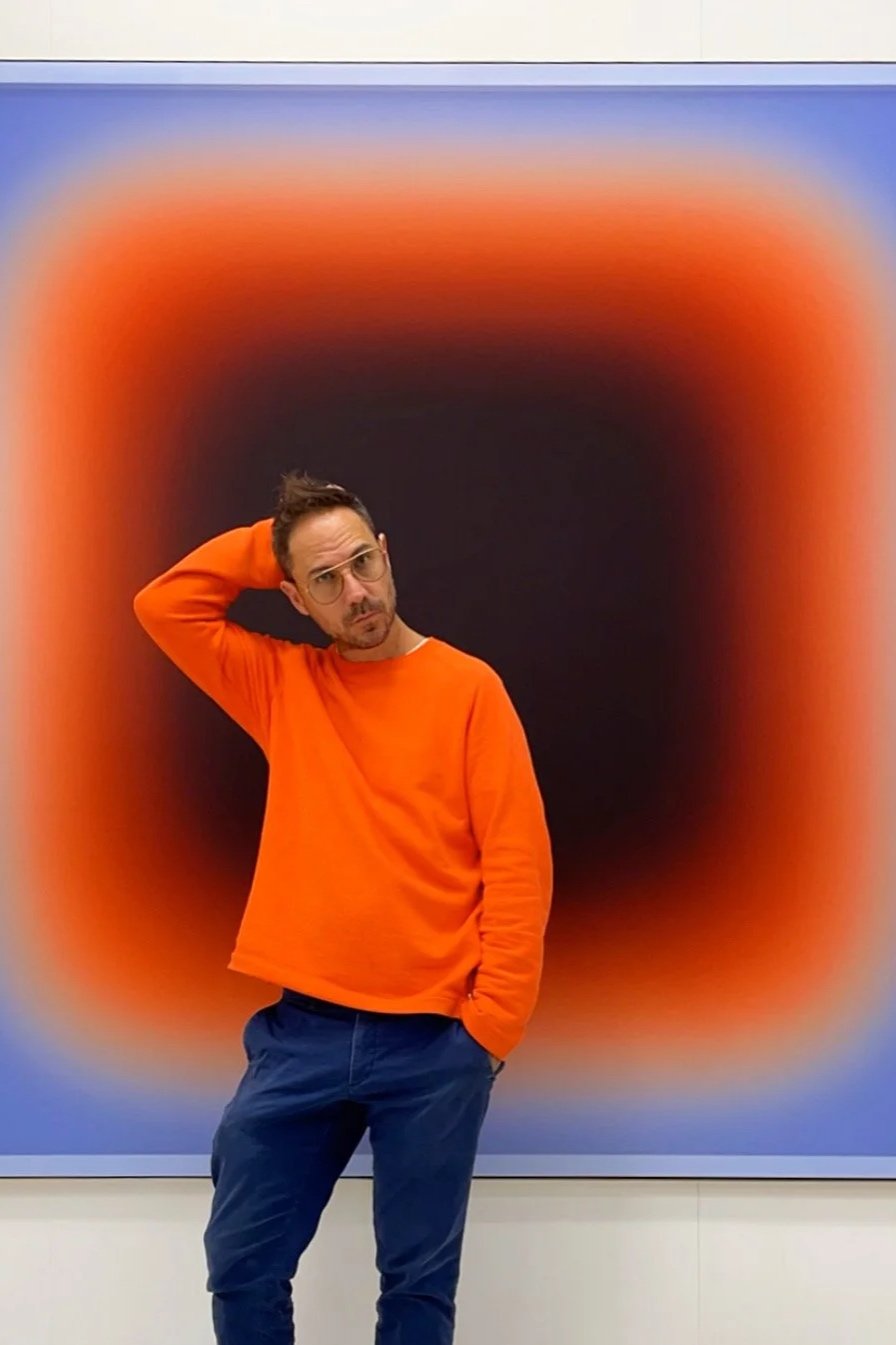 A man with glasses and a beard, wearing an orange sweater and blue pants, stands in front of an abstract art piece with a black center and a vibrant orange-red gradient border.