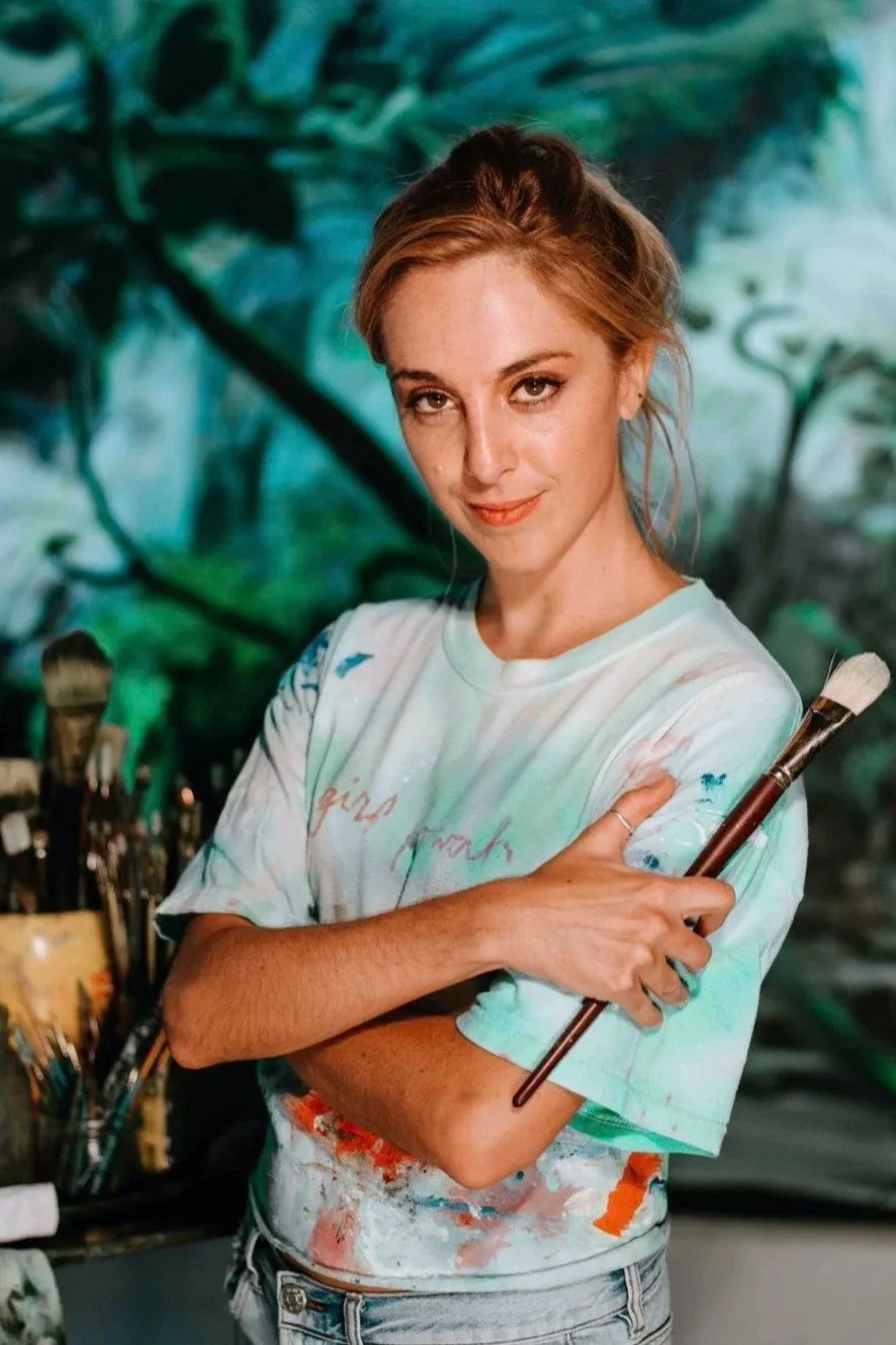 A young woman with red hair standing in front of a colorful, abstract background, holding a paintbrush. She is wearing a tie-dye t-shirt and has a confident expression.