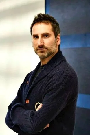 A man with dark hair and facial hair, wearing a dark cardigan, standing with arms crossed in front of a blurred background.