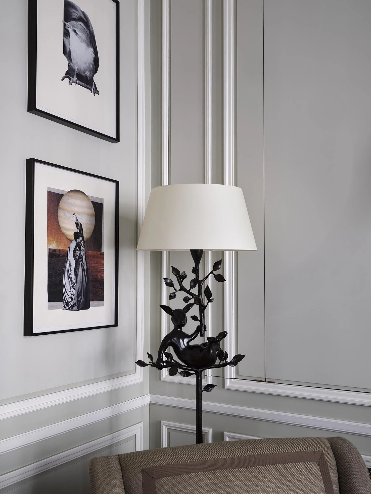 A corner of a room with framed black and white art on the wall, a white wall, and a decorative lamp with a black sculpture of a girl, a tree, and a bird on the stand, with a beige upholstered chair in the foreground.