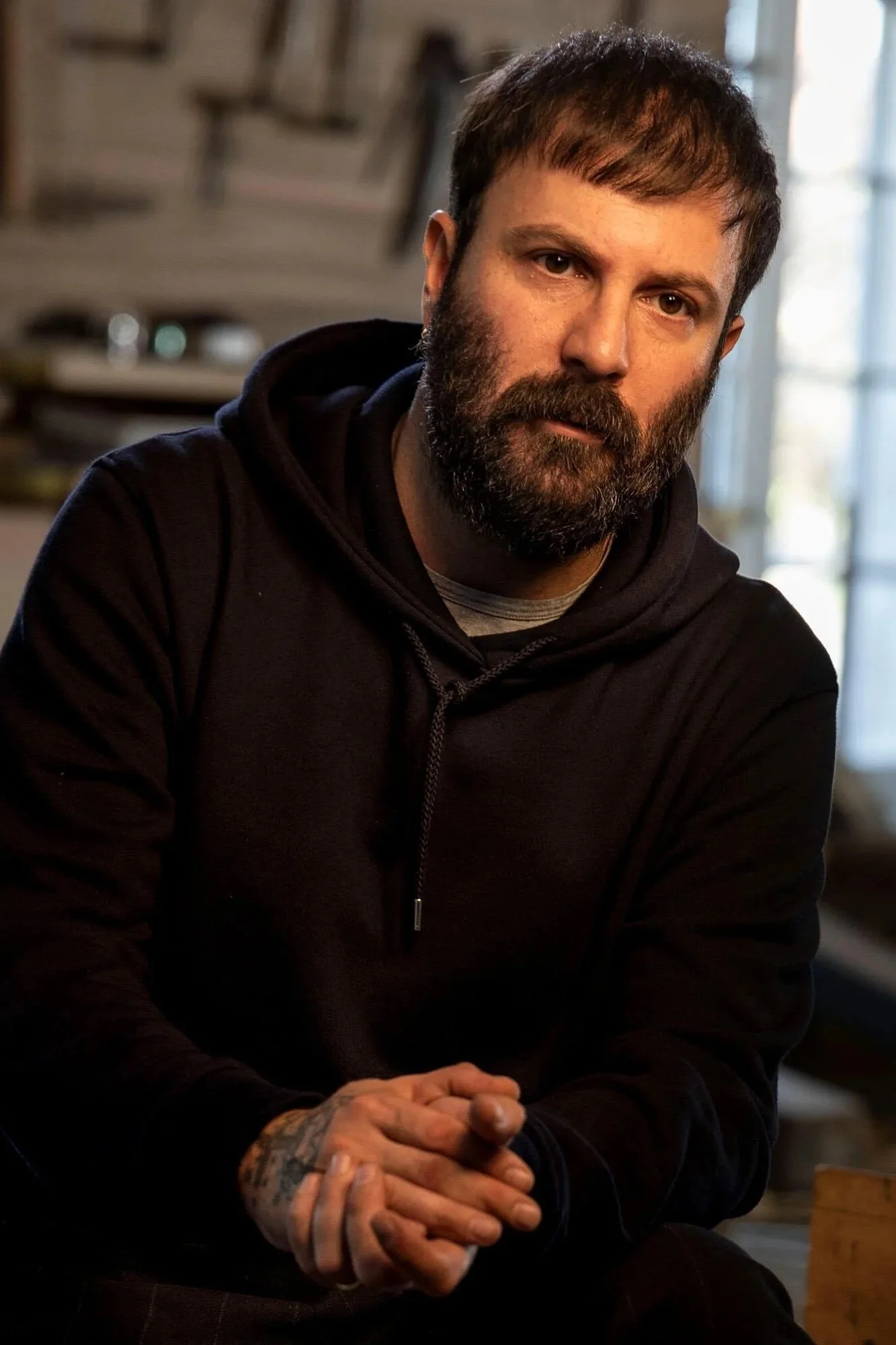 A man with a beard and short brown hair sitting indoors, wearing a black hoodie, with a tattooed hand clasped in front of him and a serious expression.