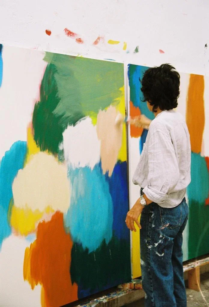 Person with curly black hair wearing a white shirt and blue jeans, painting large colorful abstract shapes on a wall in an art studio.