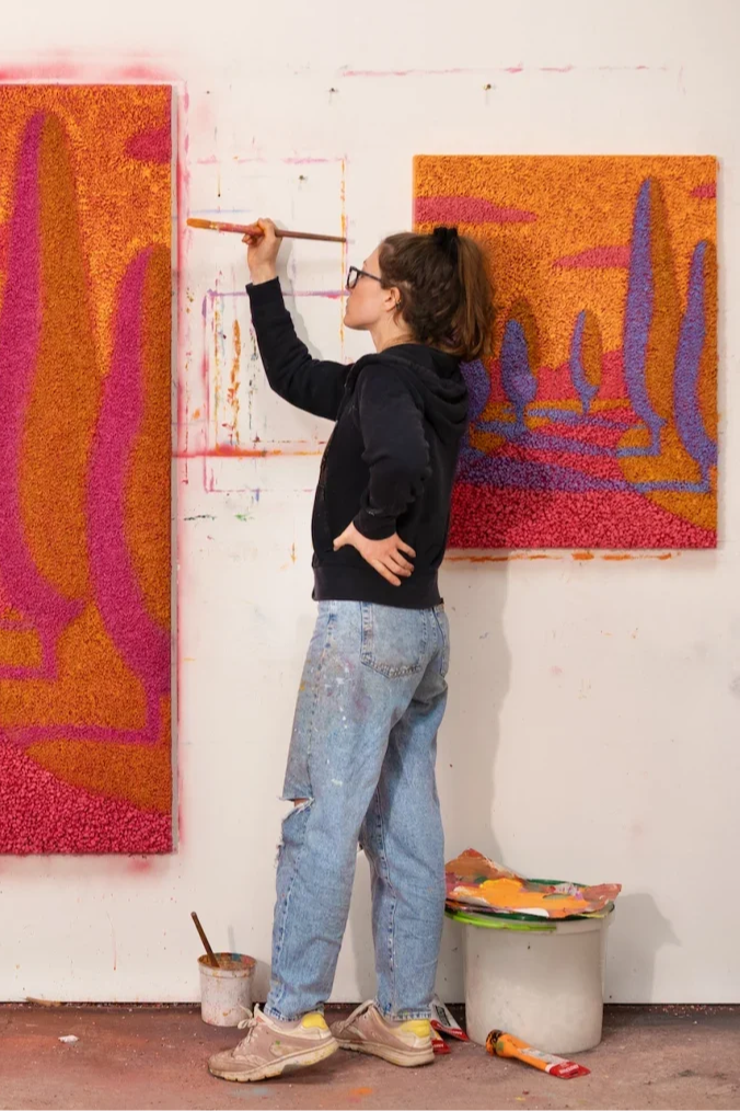 A woman with glasses and brown hair tied back, wearing a black hoodie and distressed jeans, painting with a brush on a wall in an art studio. Two colorful abstract paintings are on the wall, and painting supplies are on the floor and a nearby table.