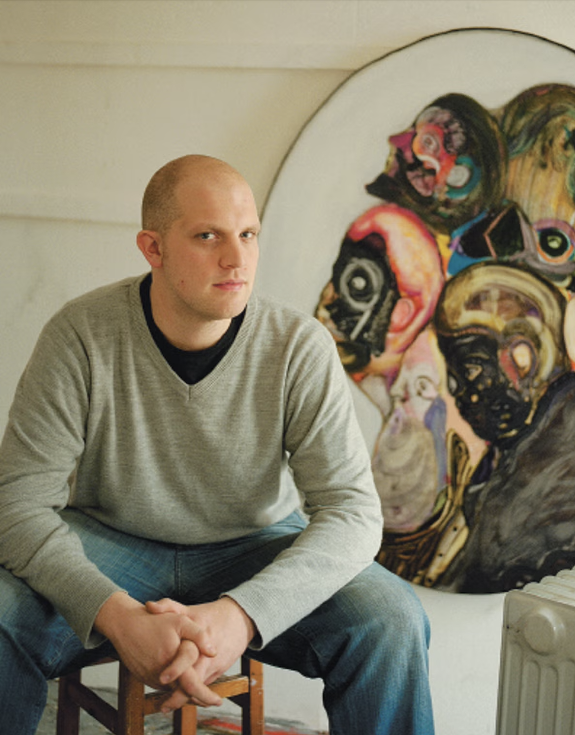 A man with a shaved head and serious expression sitting on a wooden stool in front of a colorful abstract painting.
