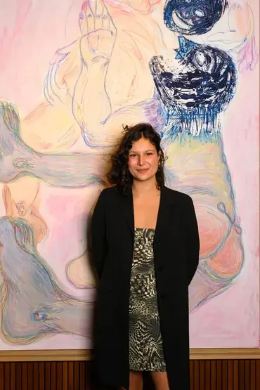 A woman with curly brown hair stands in front of an abstract pastel-colored painting with large, swirling shapes and faces. She is wearing a black coat over a patterned dress and is smiling at the camera.