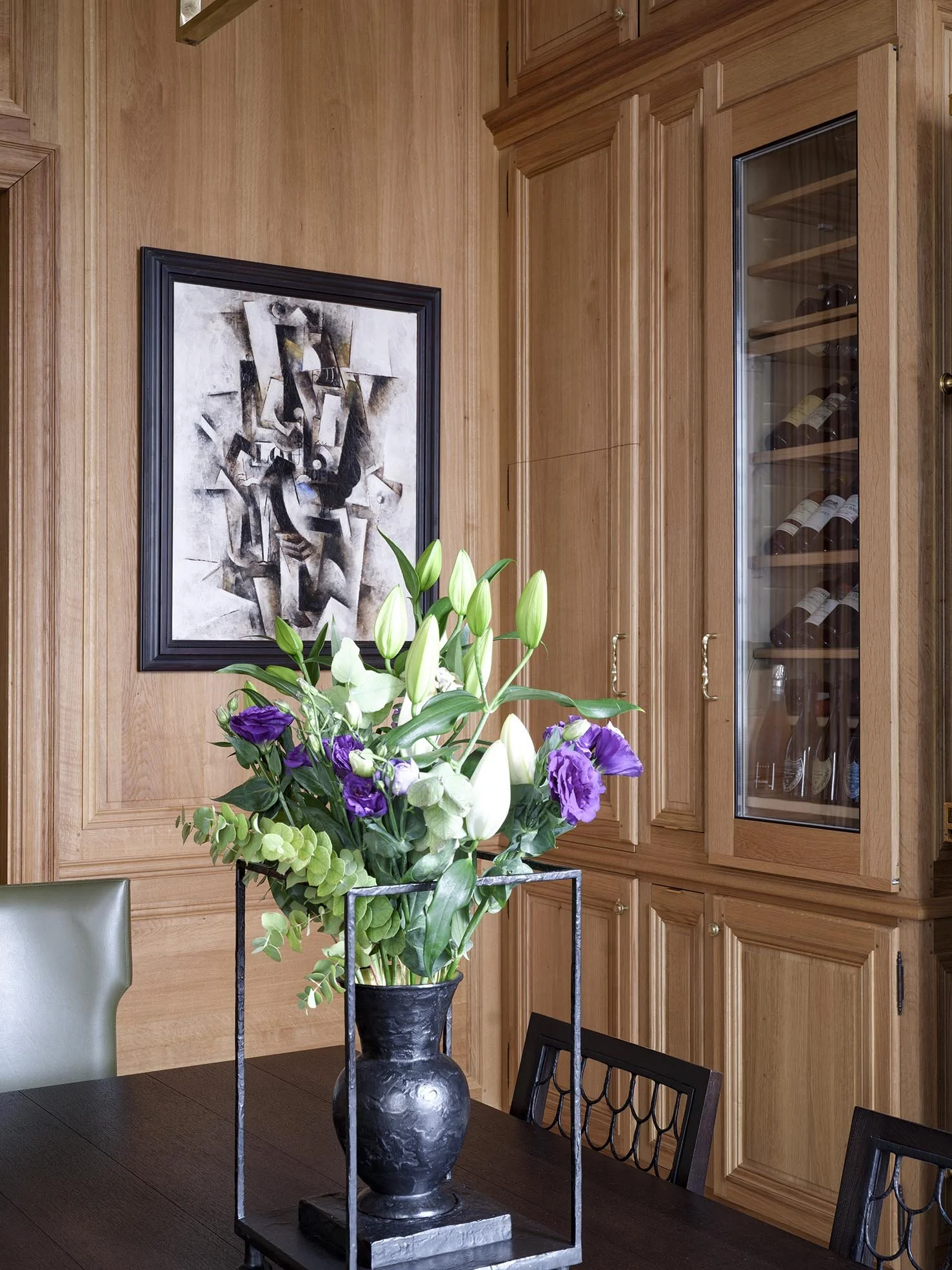 A black vase with a floral arrangement of white lilies, purple flowers, and green foliage on a dark wooden table in a room with wooden cabinetry and an abstract black-and-white painting on the wall.