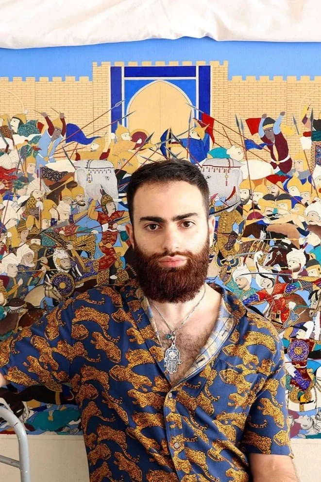 A young man with a beard and mustache taking a selfie in front of a detailed traditional-style artwork depicting a historical battle scene with soldiers, horses, and a fortress.