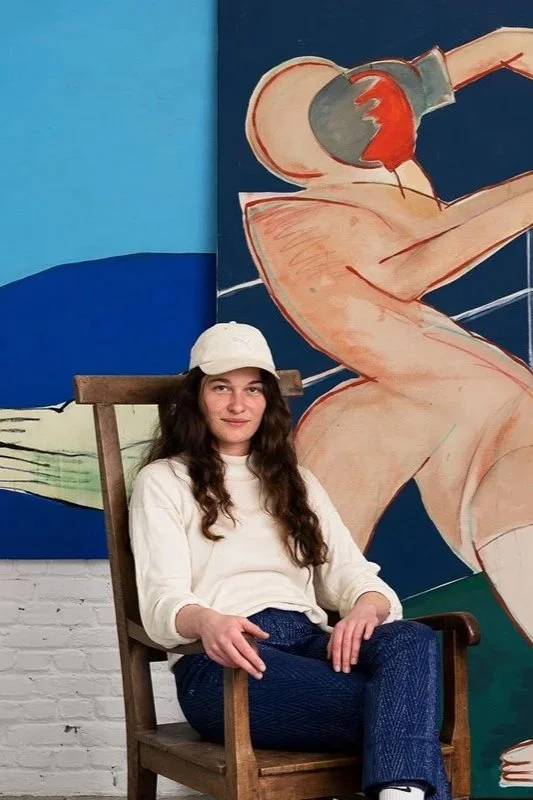 A young woman with long dark hair, wearing a white baseball cap, cream-colored sweatshirt, and blue pants, sitting on a wooden chair in front of a colorful mural of a nude figure with a football helmet.