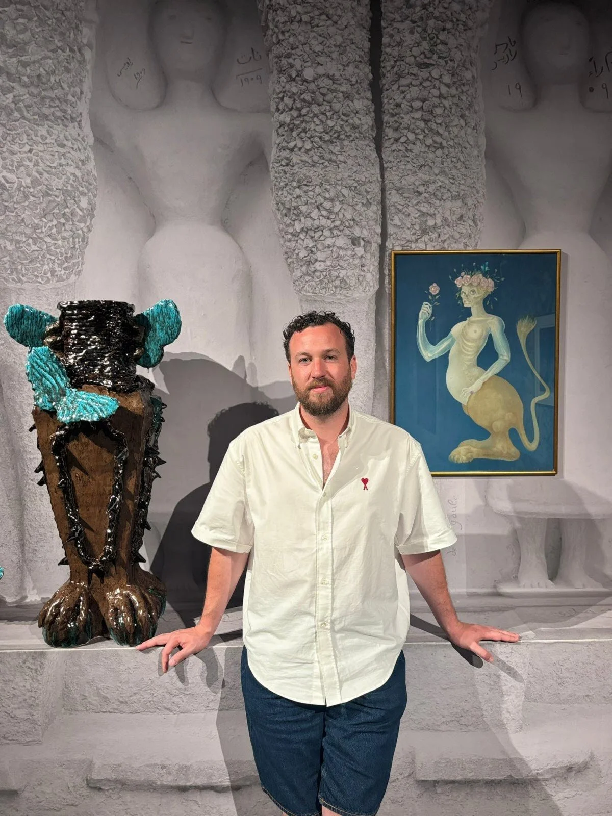 A man with a beard, wearing a cream-colored short sleeve shirt and denim shorts, standing in an art gallery with modern and surrealist artworks behind him.