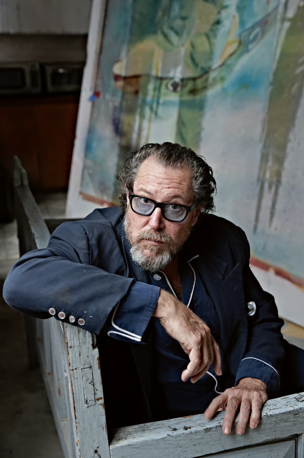 An older man with glasses, gray hair, and a beard, wearing a dark blue jacket with white piping, sitting at a rustic wooden table in an art studio. In the background, there is a large abstract painting with shades of blue, green, and yellow.