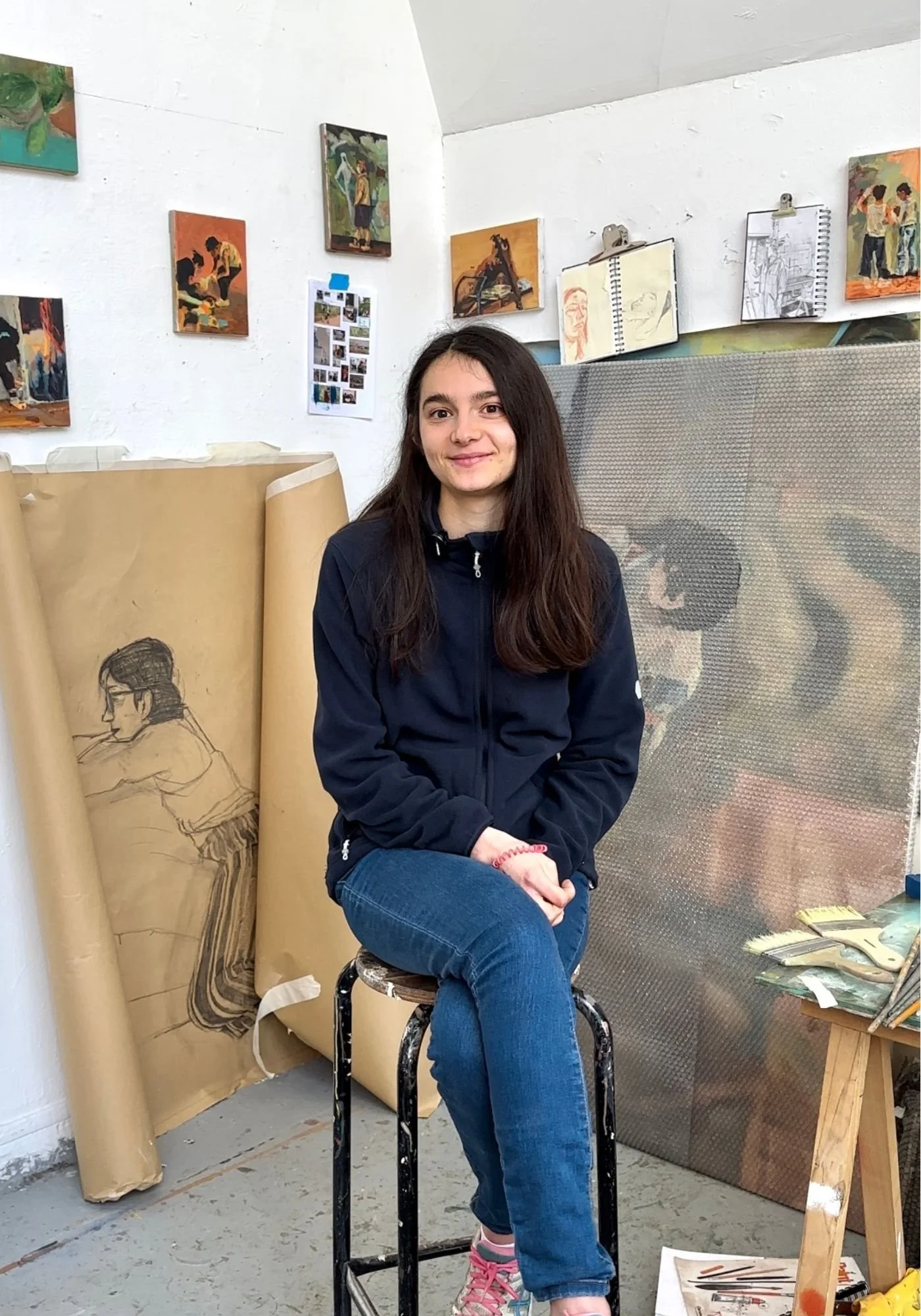 A young woman with long dark hair sits on a stool in an art studio surrounded by paintings, sketches, and art supplies.