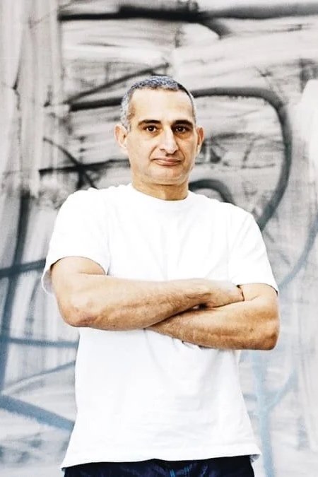 Man with short gray hair wearing a white T-shirt standing with arms crossed in front of an abstract black and white background.