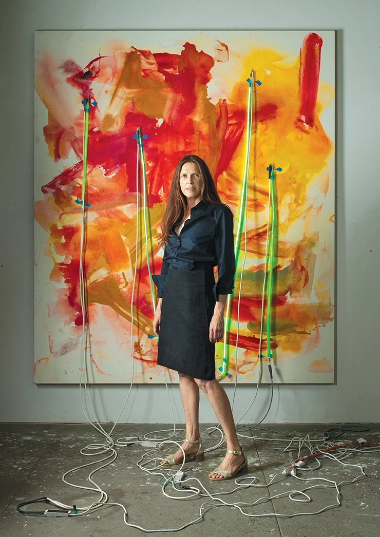 A woman standing in front of a large abstract painting with red, orange, and yellow colors, connected to neon light wires and cords.