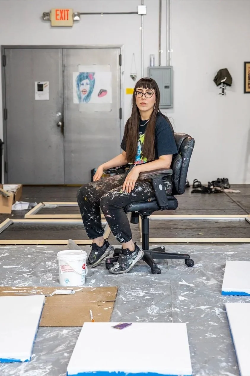 A young woman with long brown hair and glasses, wearing a black t-shirt and paint-splattered pants, sitting on a black adjustable chair in an art studio. The studio has canvases, paint supplies, and a gray door with a colorful artwork taped on it, and various equipment and supplies are visible around.