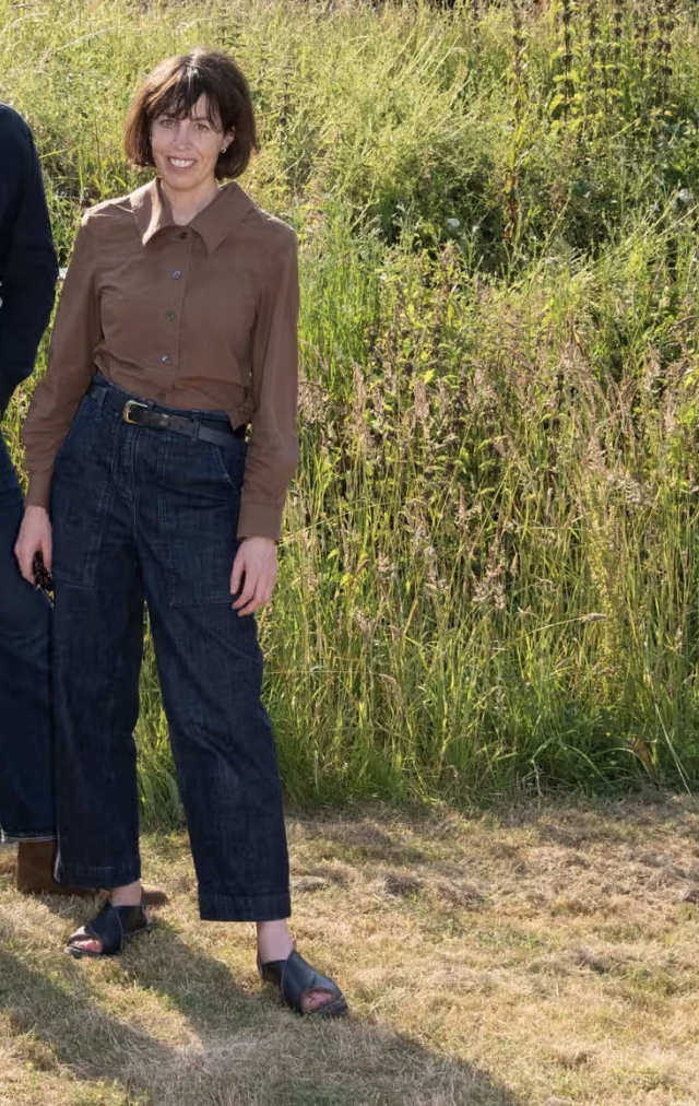 Woman with short dark hair wearing a brown button-up shirt, high-waisted dark jeans, and black sandals standing outdoors on grass with tall wild plants in the background.