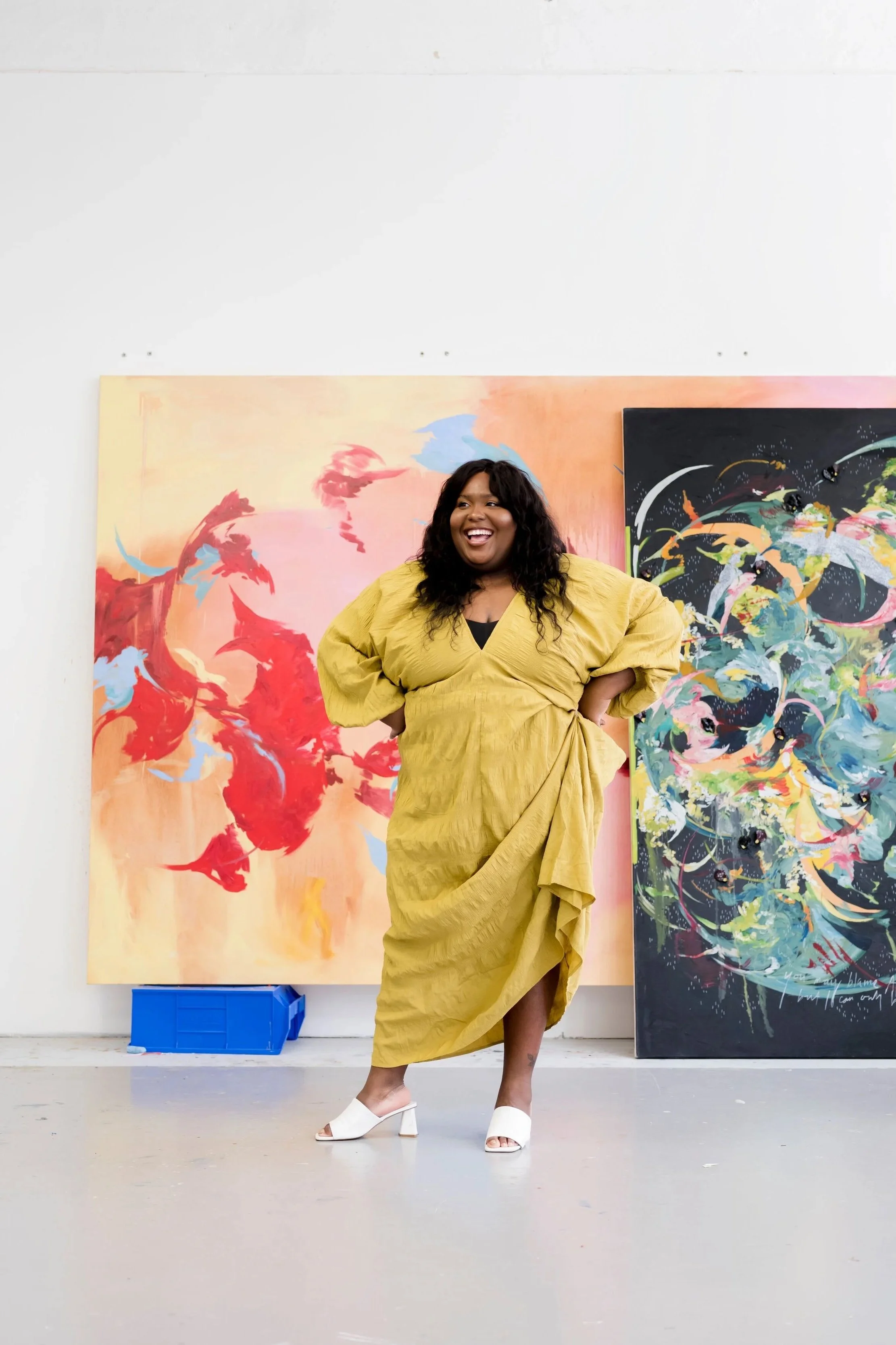 A woman with long curly black hair is standing in an art gallery, smiling, wearing a mustard-yellow dress with puffed sleeves and white open-toe heels. Behind her are two large abstract paintings, one with red, pink, and blue brushstrokes, and the other with multicolored swirling patterns on a dark background.