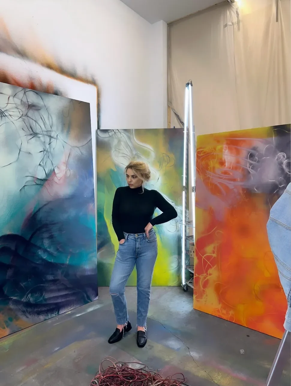 A woman with blonde hair, wearing a black turtleneck and jeans, standing in an art studio surrounded by large, abstract, colorful paintings with swirling patterns.
