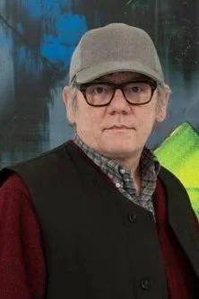 A man wearing glasses, a gray cap, a black jacket with red sleeves, and a checkered shirt stands outdoors with water and trees in the background.