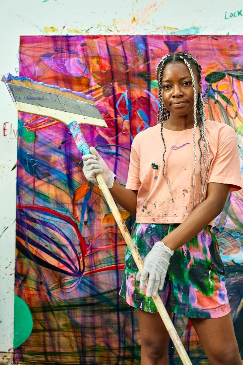 Young woman standing in front of a colorful abstract painting, holding a paintbrush and wearing paint-splattered clothes and gloves.