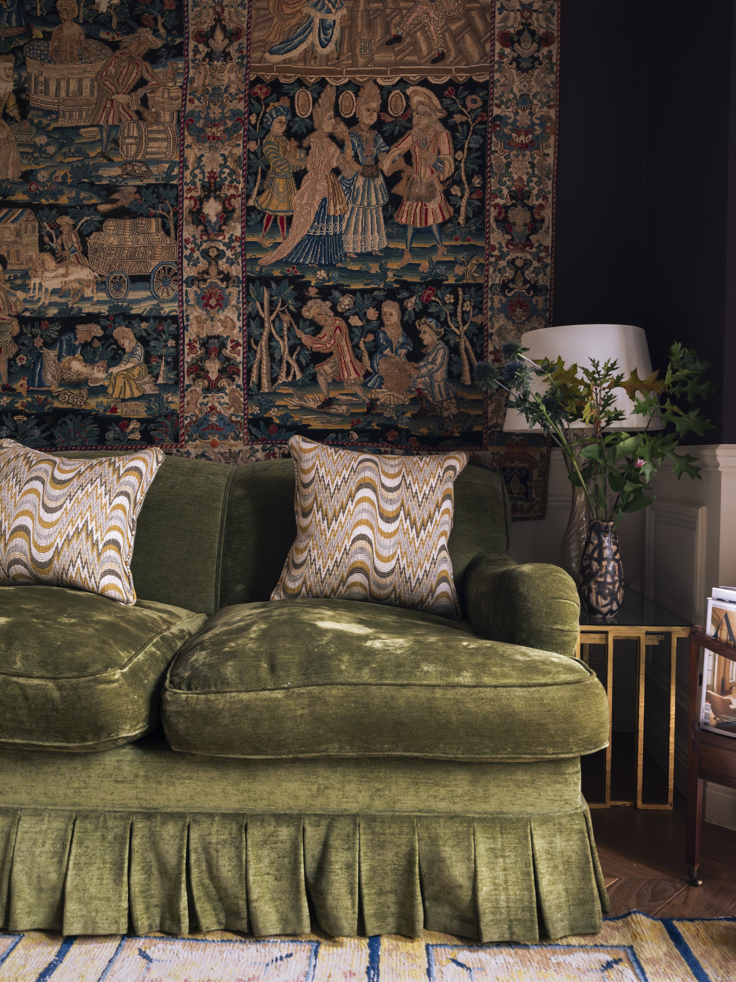 A green velvet sofa with two patterned pillows, a tapestry with intricate scenes hanging on the wall behind, a side table with a plant in a decorative vase, and a lamp beside the plant in a well-decorated living room.