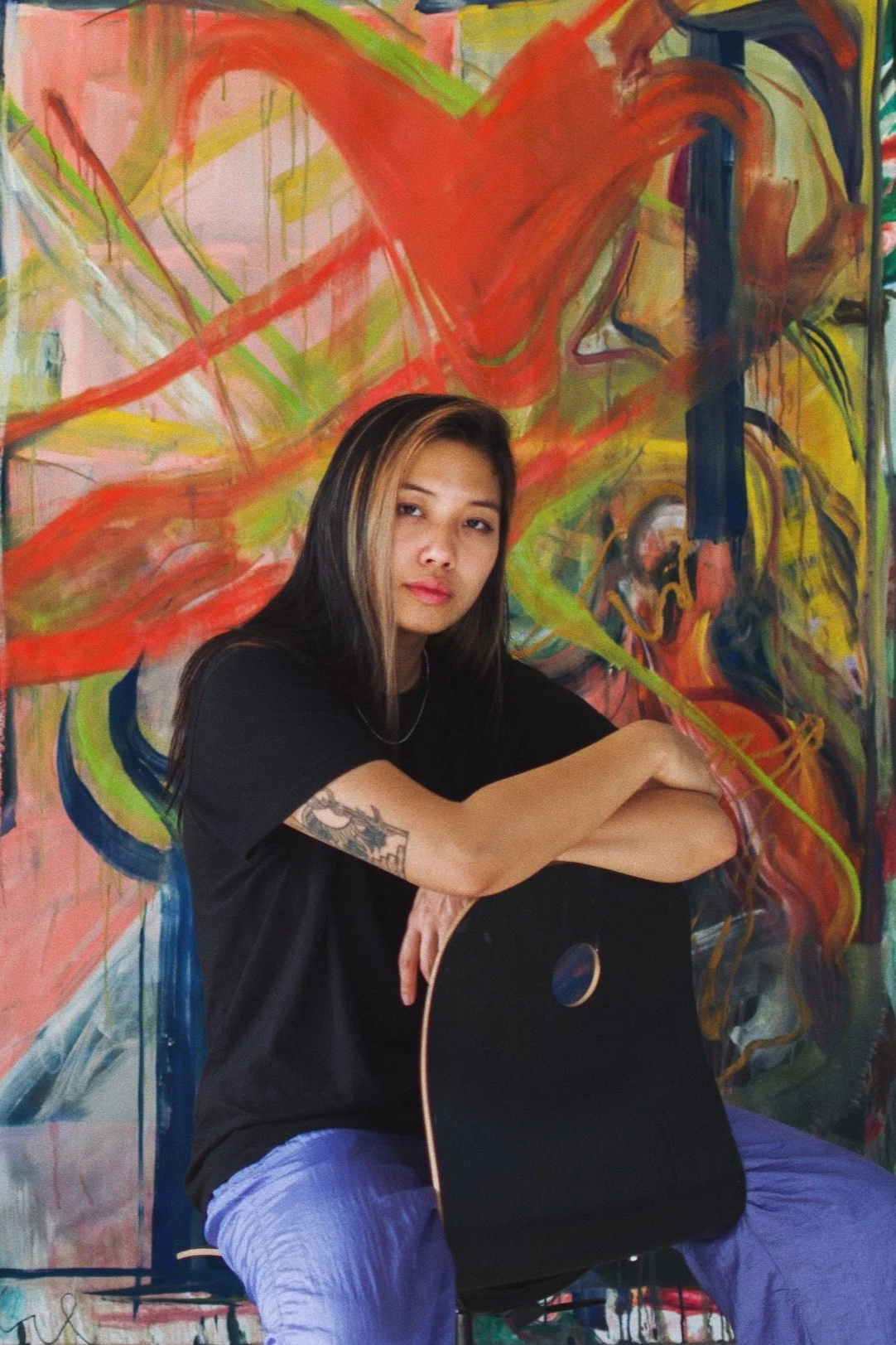 A young woman with long dark hair and tattoos, sitting on a chair with arms resting on her knees, holding an acoustic guitar, in front of a colorful abstract mural background.