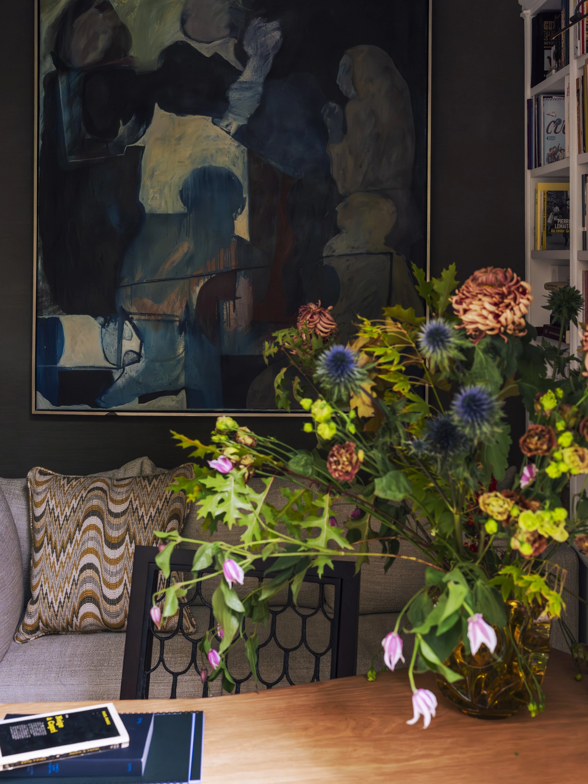 A living room with a large colorful painting on the wall, a beige sofa with patterned pillows, a wooden table with a bouquet of mixed flowers in a glass vase, and a bookshelf filled with books.