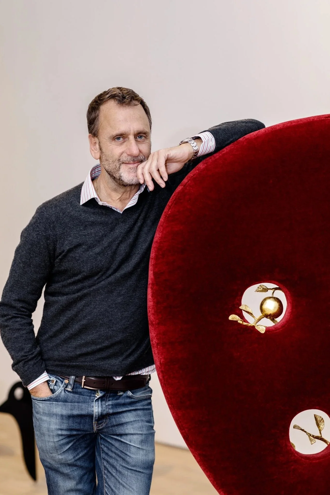 A man with a beard and short hair, wearing a dark sweater over a striped shirt, standing next to a large, red velvet heart sculpture with gold accents and circular openings.