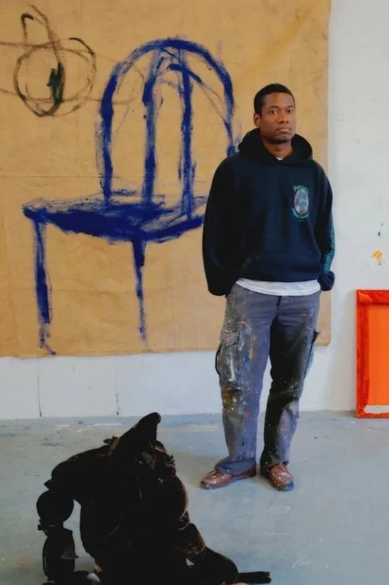 A young man standing inside an art studio, wearing a dark hoodie and paint-splattered pants, with a black dog sitting on the concrete floor nearby. Behind him, a large canvas features a blue outline of a chair and a small circle with abstract markings, with a white wall to the side.