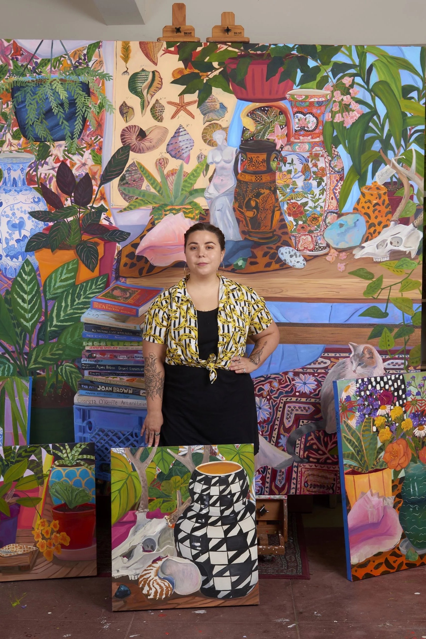 A woman stands in front of a vibrant, colorful still life painting featuring various vases, plants, shells, and animals, including a cat on a table to her right, with additional paintings of plants and objects on the floor.