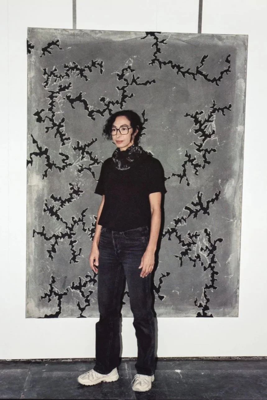 A woman with curly dark hair, wearing glasses, a black T-shirt, dark jeans, white shoes, and a patterned scarf around her neck, standing in front of an abstract black and gray painting with jagged, branch-like designs.