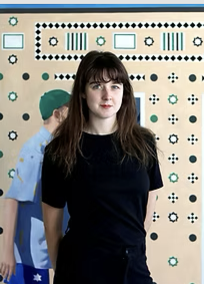 Young woman with long brown hair wearing a black shirt standing indoors in front of a colorful, patterned wall.