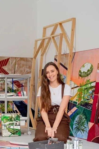 Young woman with long hair smiling in an art studio, surrounded by wooden frames and colorful paintings.