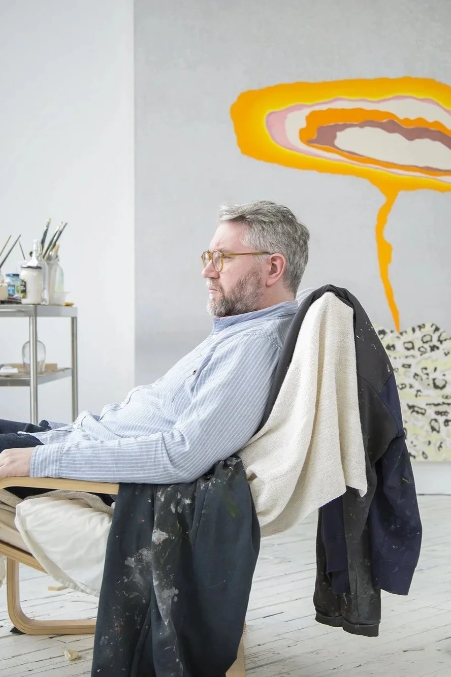 A man with gray hair and glasses sitting in an art studio. He is wearing a light blue striped shirt and has a white towel and painter's jacket draped over his chair. Behind him, there is a colorful abstract painting on the wall, and to the side, a small cart with art supplies.