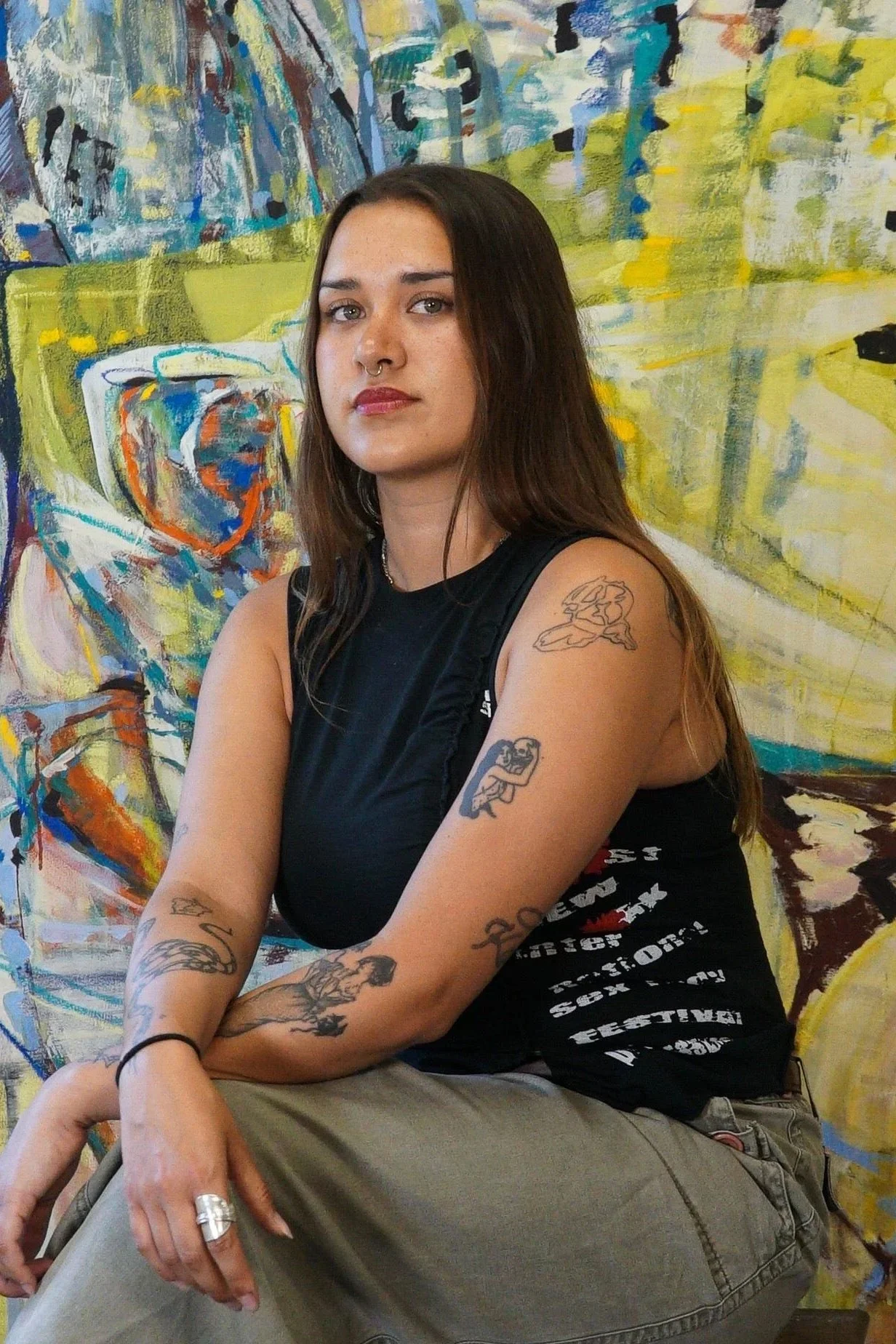 A young woman with long brown hair and tattoos sitting in front of an abstract painting with yellow, blue, white, and orange colors.
