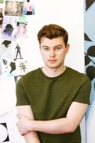 Young man with brown hair and green shirt standing with arms crossed in front of a wall with various posters and artwork
