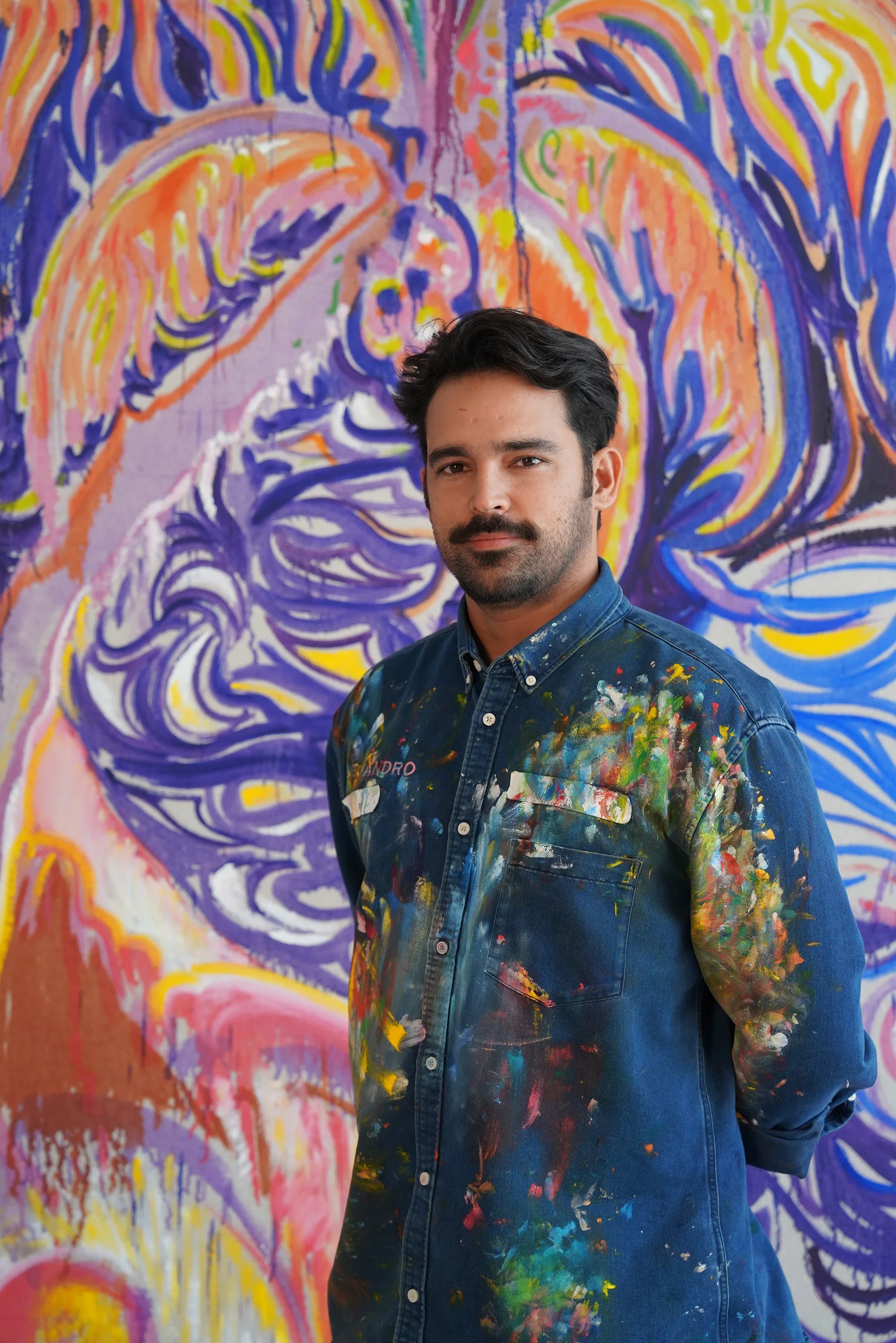 An artist stands in front of a colorful abstract painting, wearing a paint-splattered denim shirt and looking at the camera.