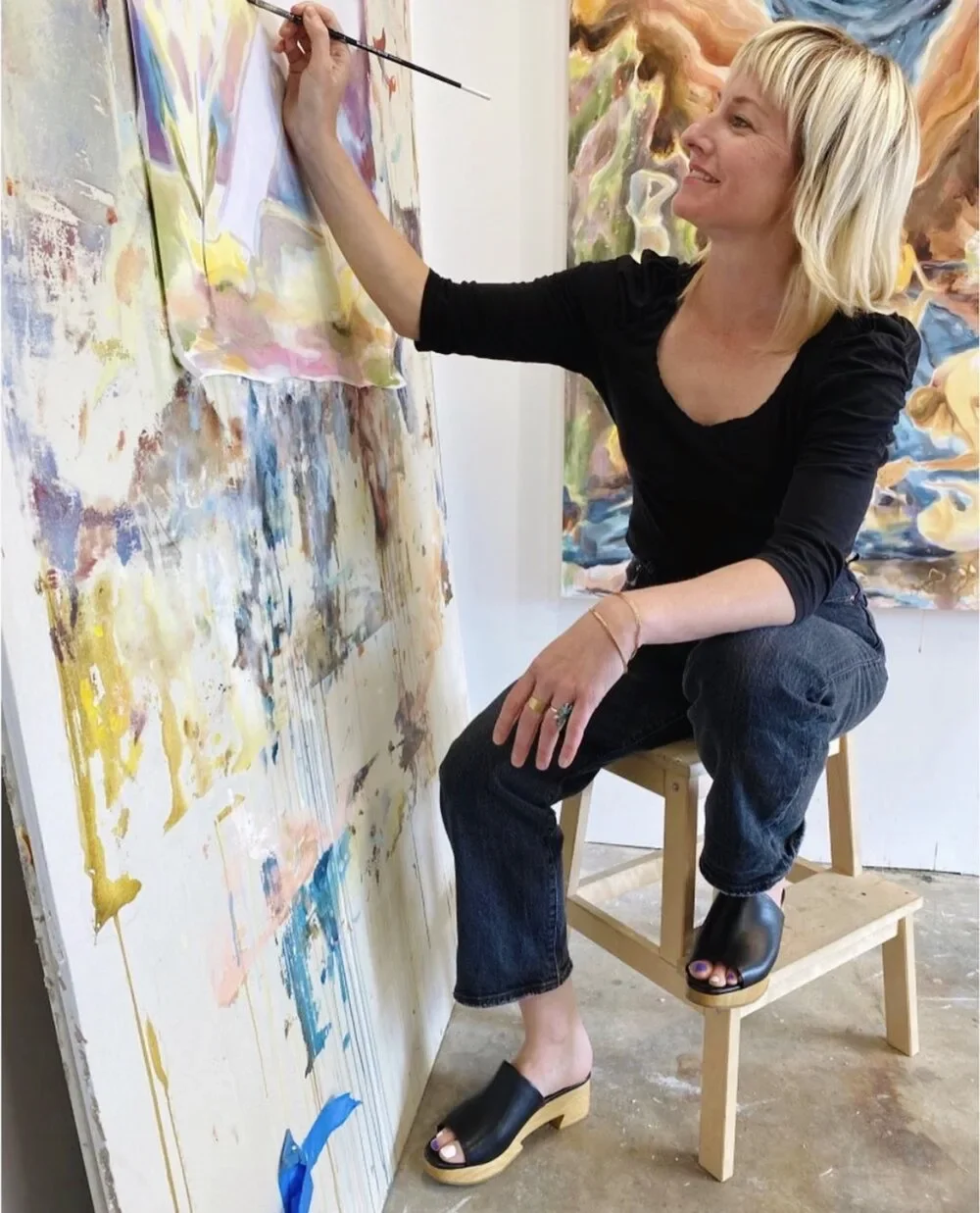 A woman with blonde hair, wearing a black top and jeans, is sitting on a stool and painting on a large canvas with colorful abstract patterns in an art studio.