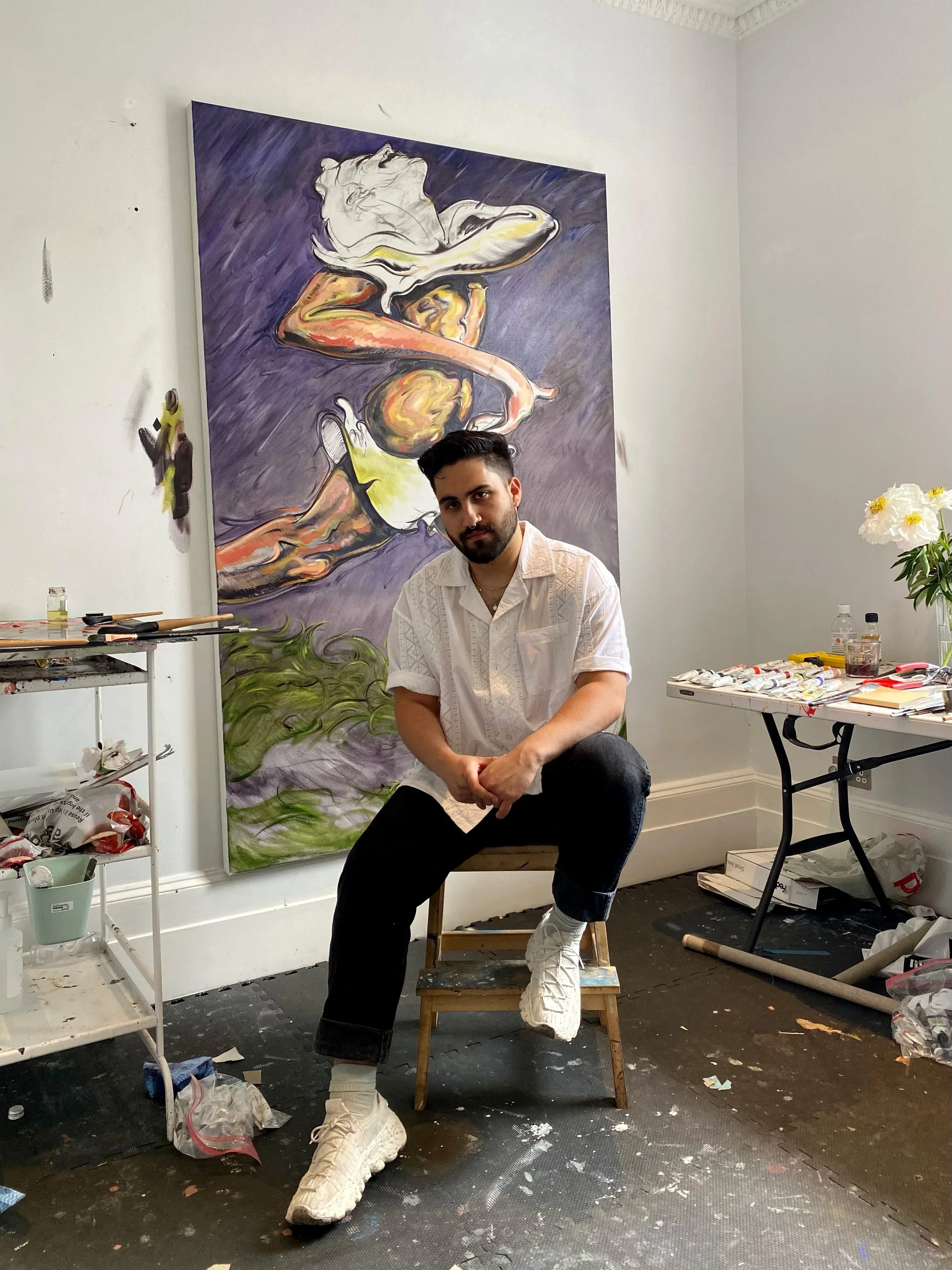 A man with dark hair and a beard, wearing a white shirt, black pants, and white shoes, sitting on a small wooden chair in an art studio. Behind him is a large, colorful painting of a female figure with white hair and a yellow skirt. There are art supplies and tools on nearby tables, and the floor has paint splatters.