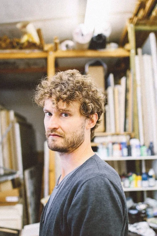 A young man with curly hair and a beard standing in an art studio or workshop with shelves filled with art supplies, books, and materials in the background.