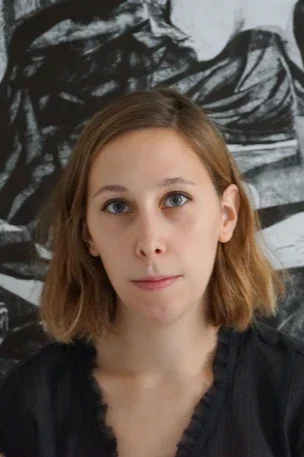 A woman with shoulder-length light brown hair and blue eyes, wearing a black top, standing in front of a black and white abstract background.