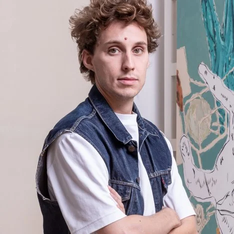 A young man with curly hair and a light complexion standing with arms crossed, wearing a white shirt and a blue denim vest, next to a colorful abstract painting.