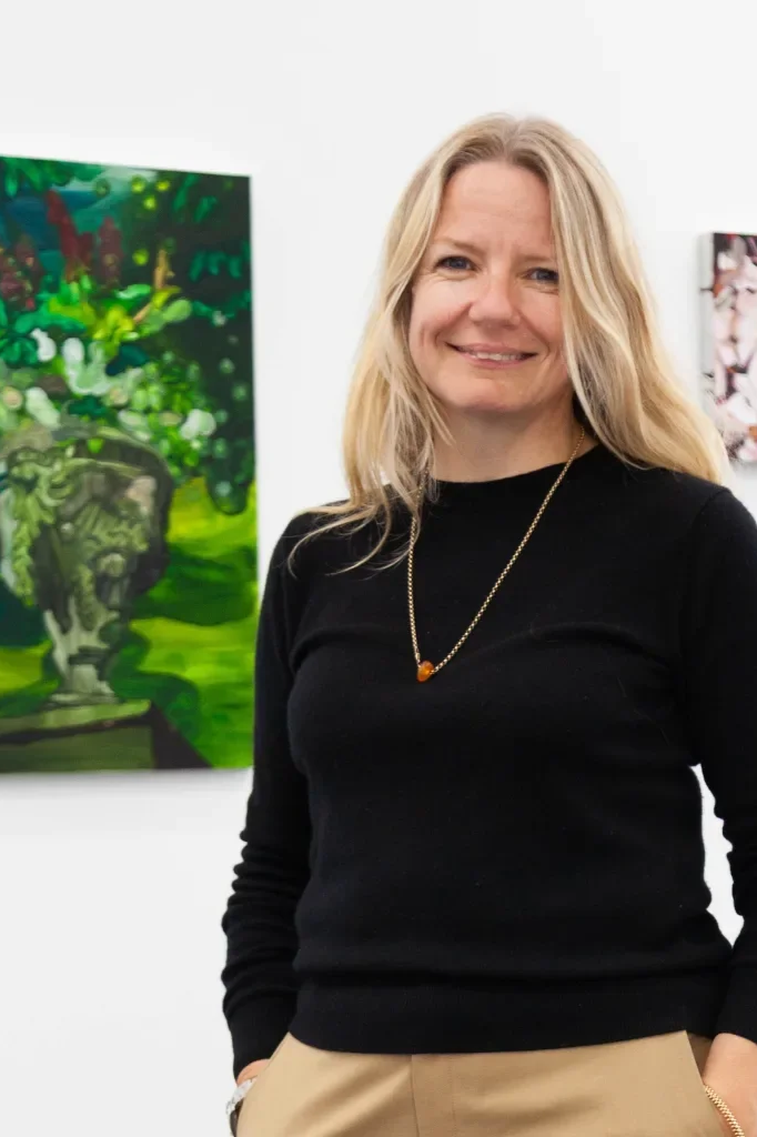 A woman with blonde hair smiling, wearing a black sweater and beige pants, stands in front of colorful paintings on a white wall.