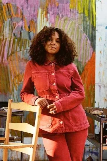 Young woman with curly hair standing in front of a colorful abstract mural in an art studio, wearing a red jacket and matching pants.