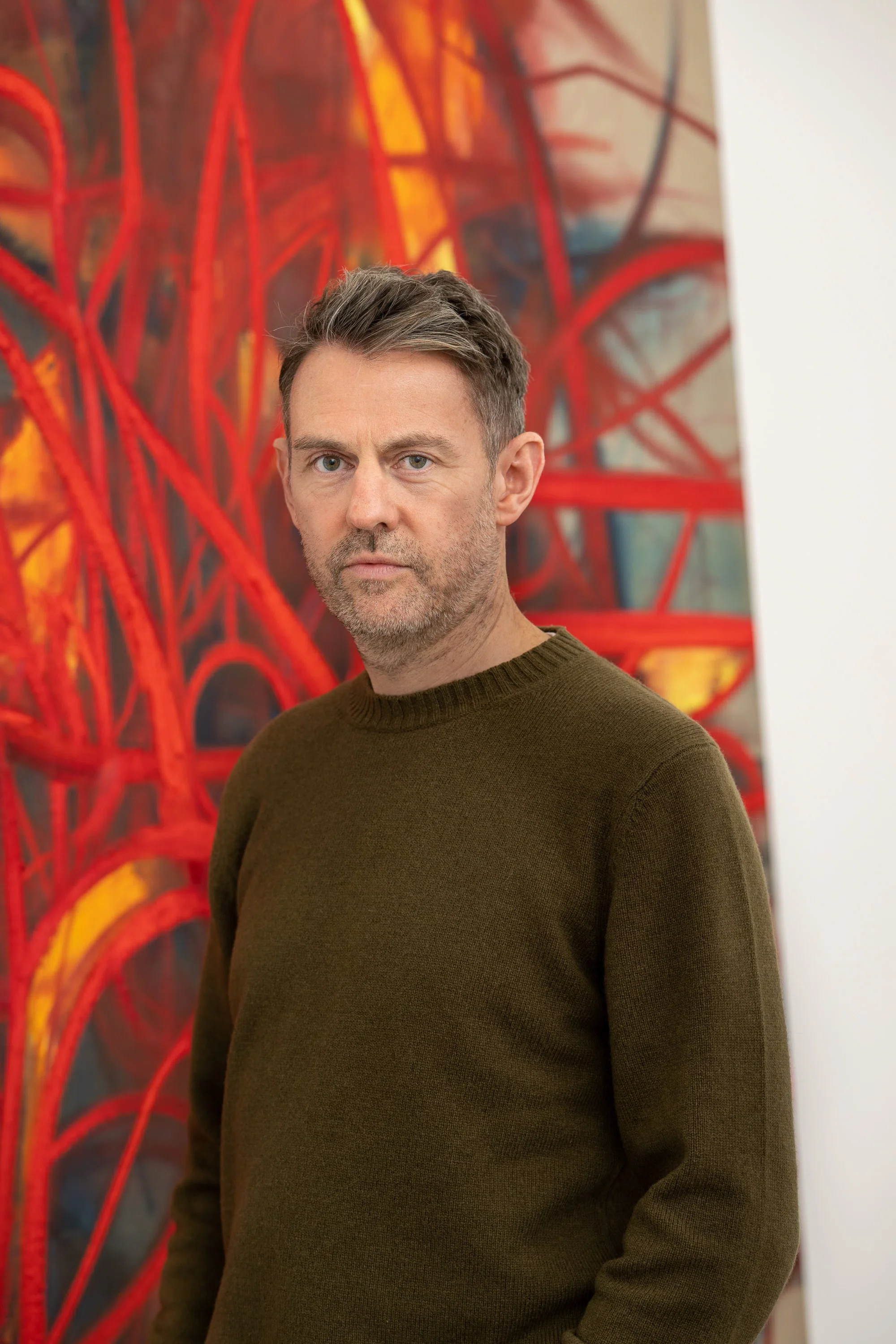 A man with short brown hair and a beard wearing a brown sweater standing in front of an abstract colorful painting with red, yellow, and blue hues.