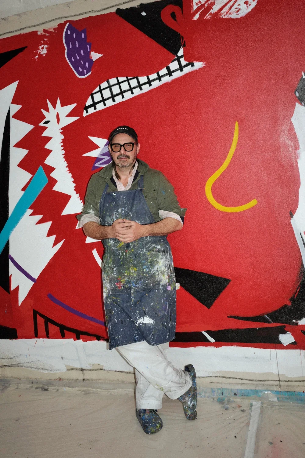 An artist stands in front of a large, colorful abstract mural featuring sharp angles, bold lines, and bright red, black, white, purple, yellow, and blue colors. The artist is wearing paint-splattered clothes, including a dark apron and a cap, and has his hands clasped in front of him.