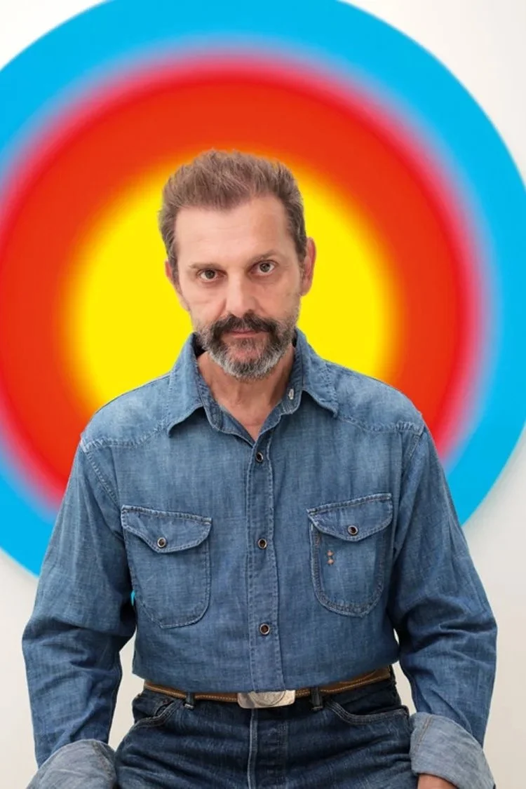 A man with a beard and mustache, wearing a denim shirt and jeans, sitting in front of a colorful circular artwork with red, yellow, and blue concentric rings.