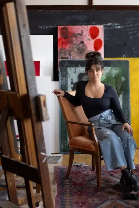A woman sitting on a wooden chair in an art studio, surrounded by paintings and an easel.